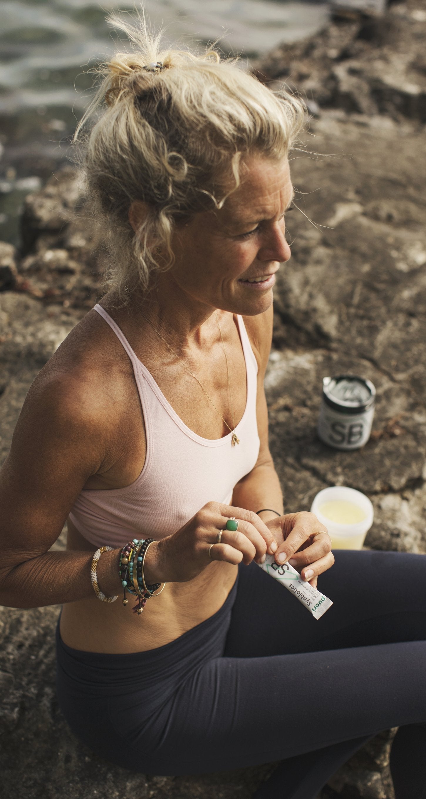 Woman sitting on rocks by the water holding a Puori supplement stick sachet, with a Puori SB tub and a glass of liquid beside her.